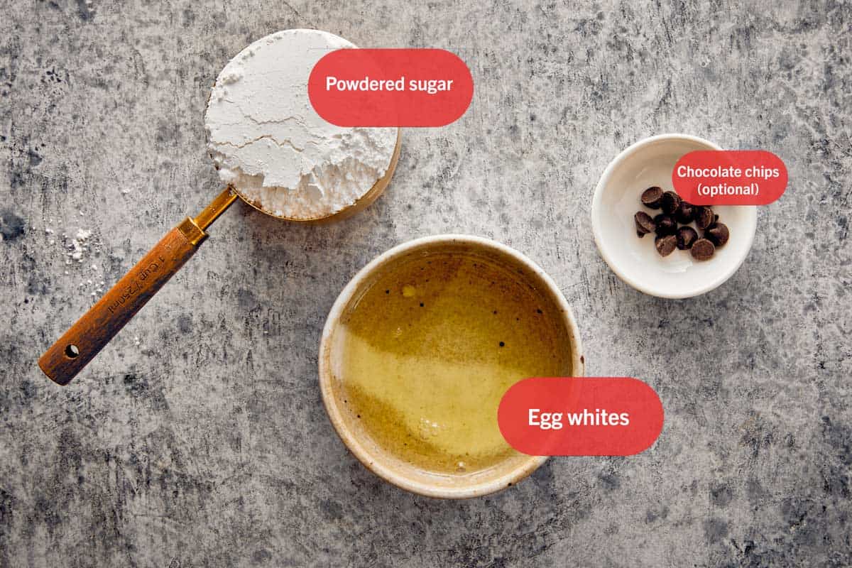 Ingredients for Swiss Meringue Ghosts including powdered sugar, egg whites, and chocolate chips labeled on a gray surface.