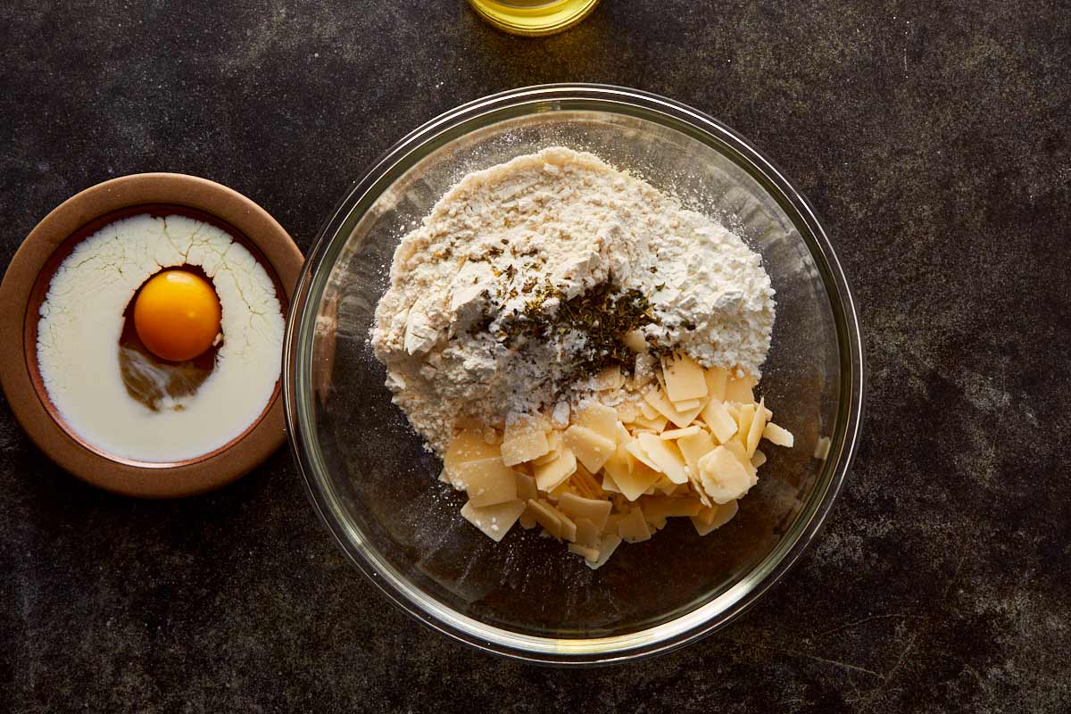 Overhead shot of flour, cheese, herbs, and milk with an egg — ingredients coming together for the grissini dough.