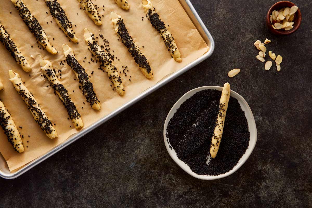 Fingers of dough being coated with black sesame seeds on a parchment-lined tray for texture and color.
