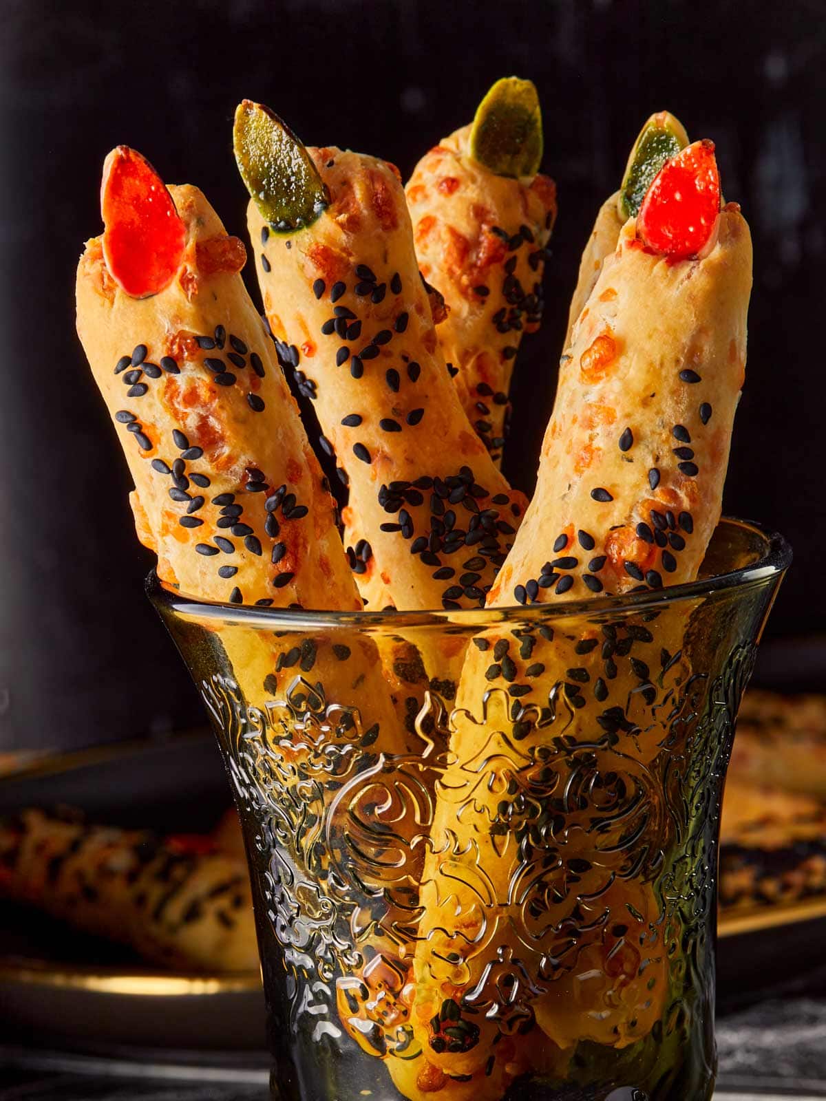 Close-up of Witch Finger Grissini breadsticks standing in a decorative glass, topped with red and green almond “nails” and coated with black sesame seeds for a spooky Halloween look.