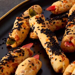 Close-up of freshly baked Witch Finger Grissini on a dark serving plate, coated with black sesame seeds and topped with red and green almond “nails” for a spooky Halloween look.