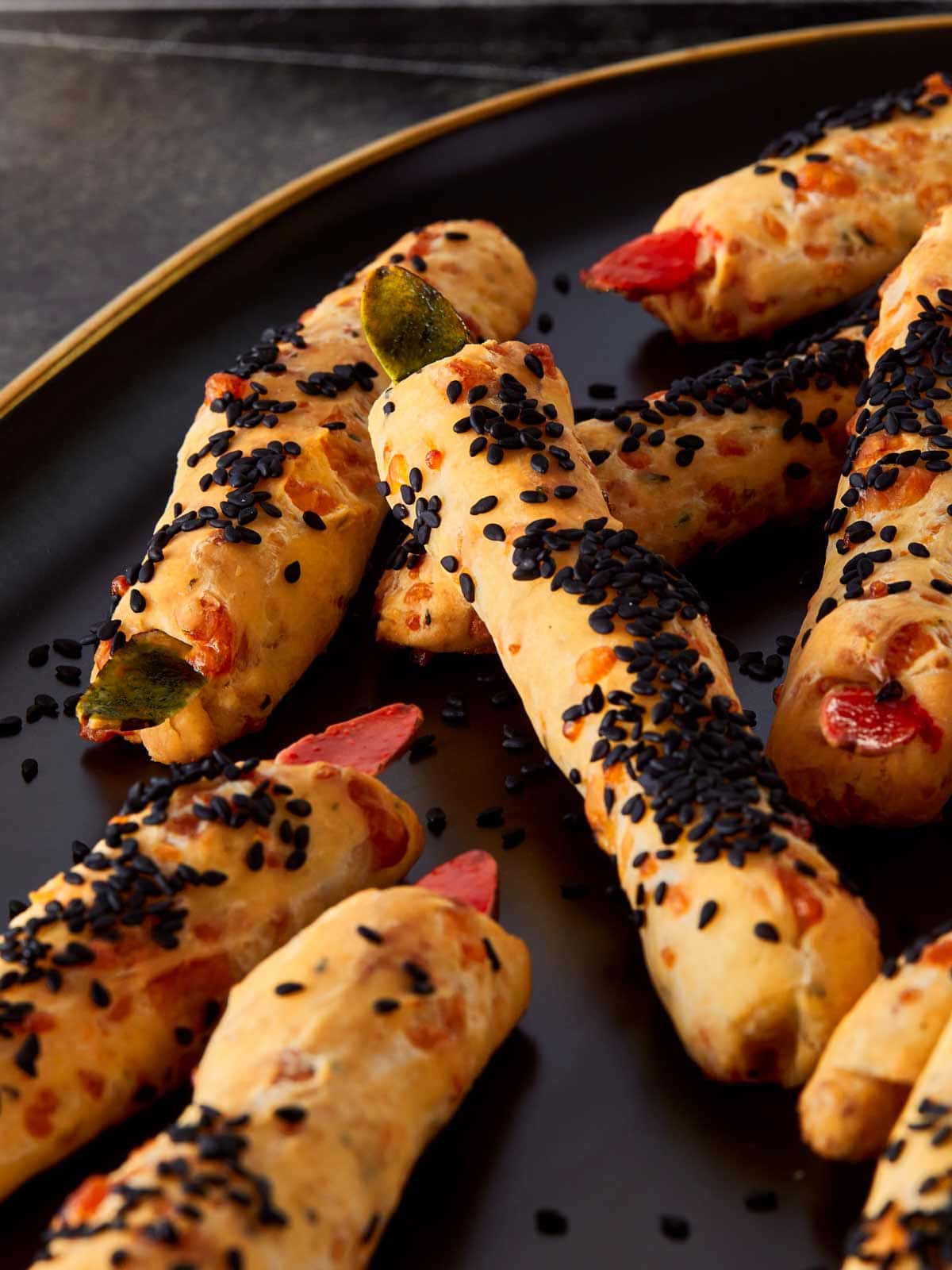 Close-up of freshly baked Witch Finger Grissini on a dark serving plate, coated with black sesame seeds and topped with red and green almond “nails” for a spooky Halloween look.