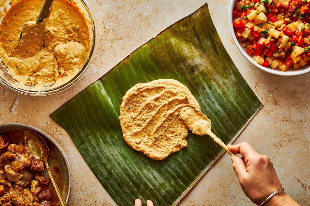 spooning the mix of meat on top of a tamales paste which is sitting on top of a green leaf from a banana tree
