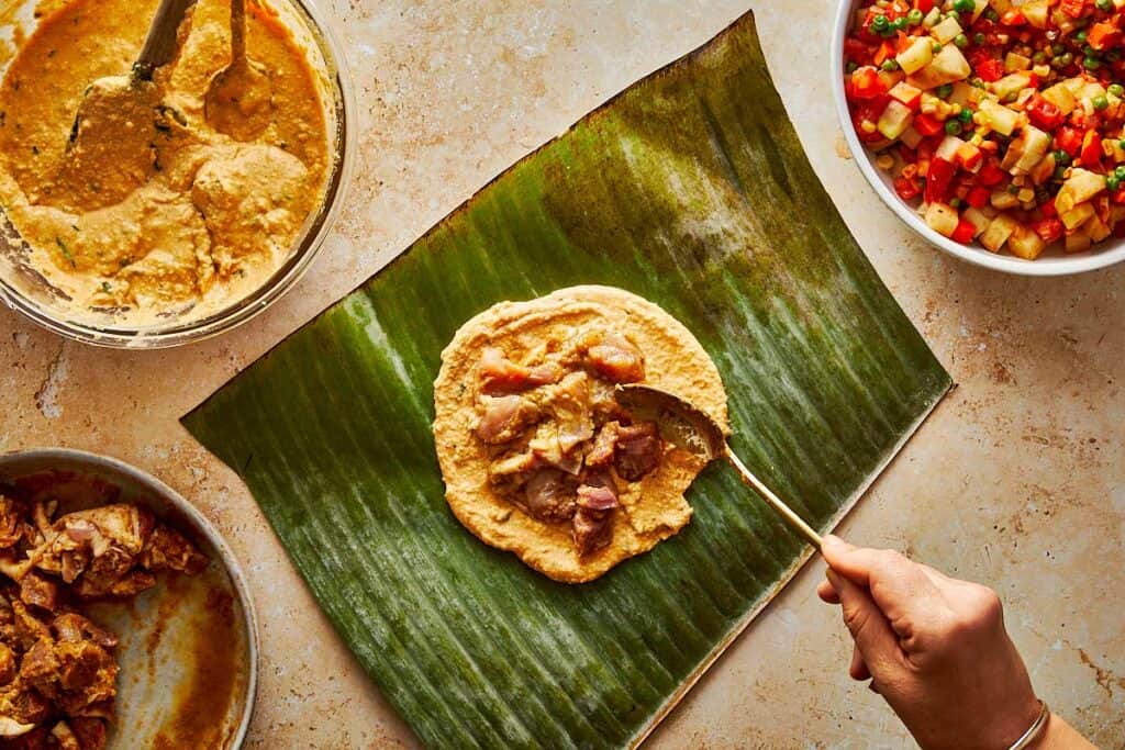 spooning the mix of meat on top of a tamales paste which is sitting on top of a green leaf from a banana tree