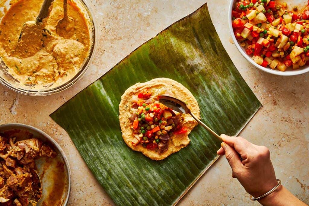 spooning the mix of meat on top of a tamales paste which is sitting on top of a green leaf from a banana tree