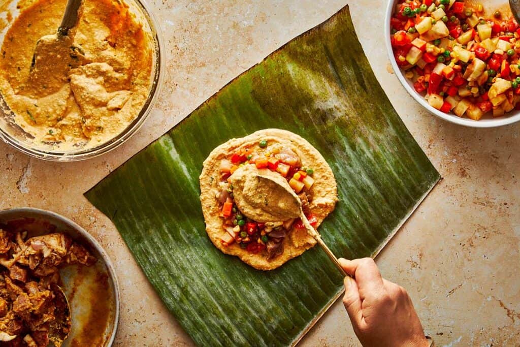 spooning the mix of meat on top of a tamales paste which is sitting on top of a green leaf from a banana tree