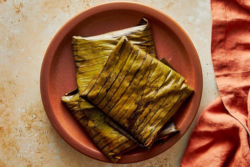 Top-down view of three square tamales wrapped in green and brown banana leaves, stacked on a terracotta-colored plate