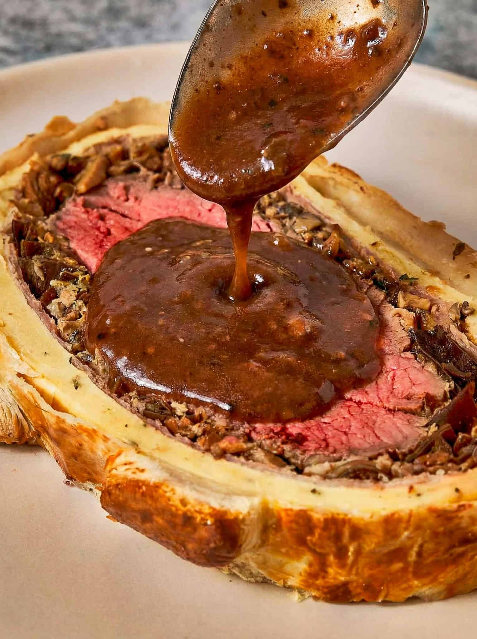 Beef wellington slice on a white plate with a special sauce of red wine poured over it with a spoon.