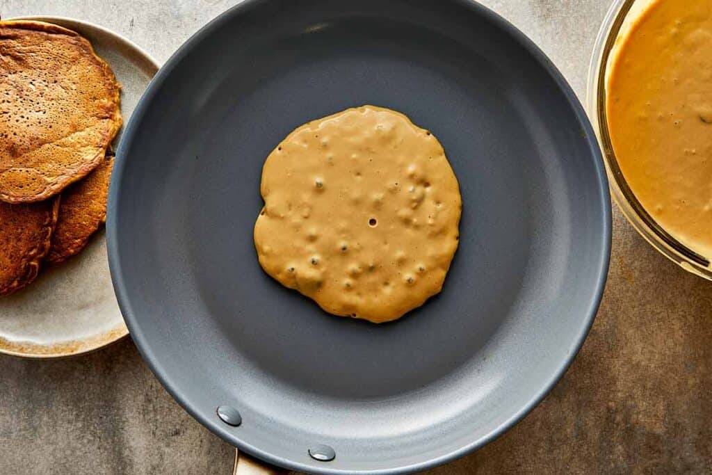 a top down shot of a pancake with the raw side up forming bubbles on the batter frying in a pan