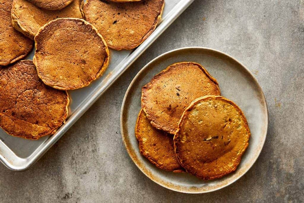 a top down shot of 3 pancakes on a plate and more pancakes sitting on a side tray