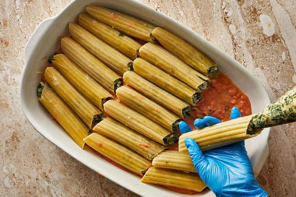 a picture of a blue glove over a hand which is stuffing cannelloni tubes with a cheese filling laying on top of the bed of sauce in a baking dish