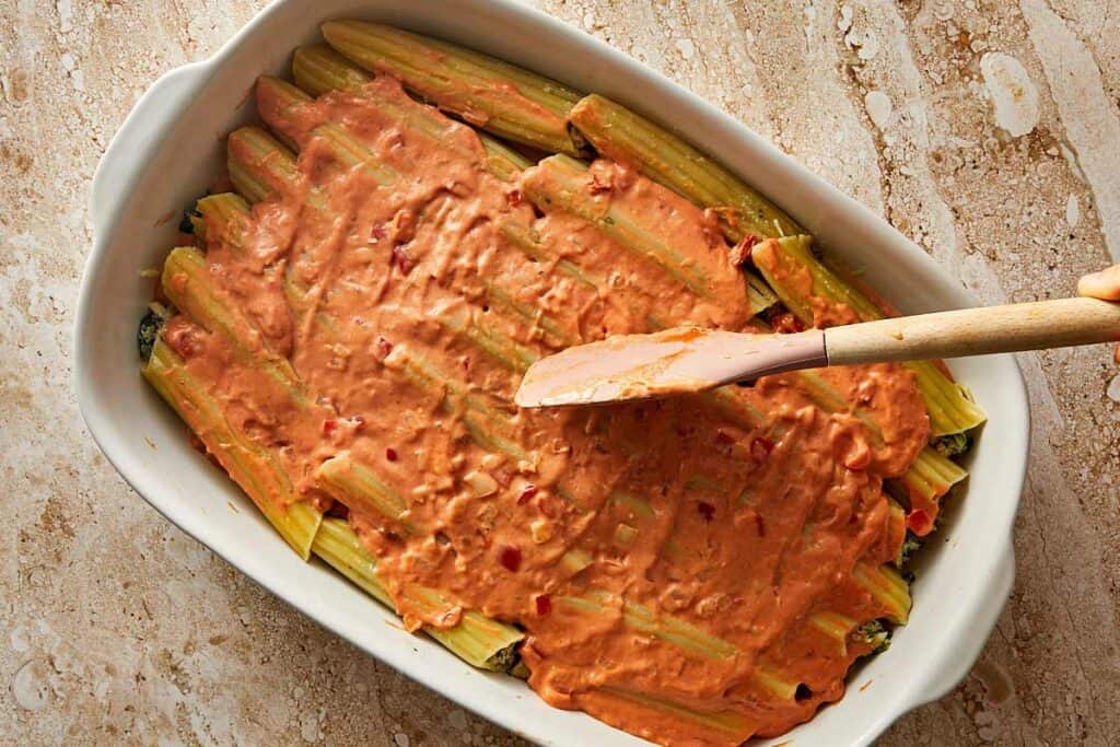 a top down shot of a full layer of cannelloni that is in a baking dish with a thick orange sauce covering it with a wooden spoon.