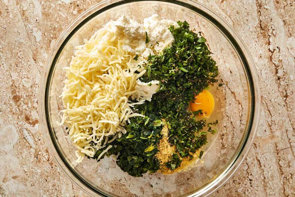 a glass mixing bowl sitting on a bench with cheese, green herbs and eggs