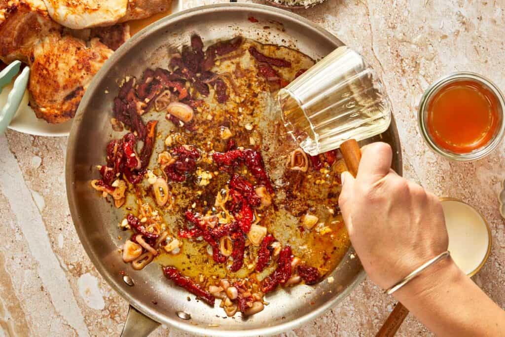 a top down shot of a bowl with many spices, sun-dried tomatoes and brown spices all mixed up, with a cup of a what looks like wine being poured into it. The cup is clear