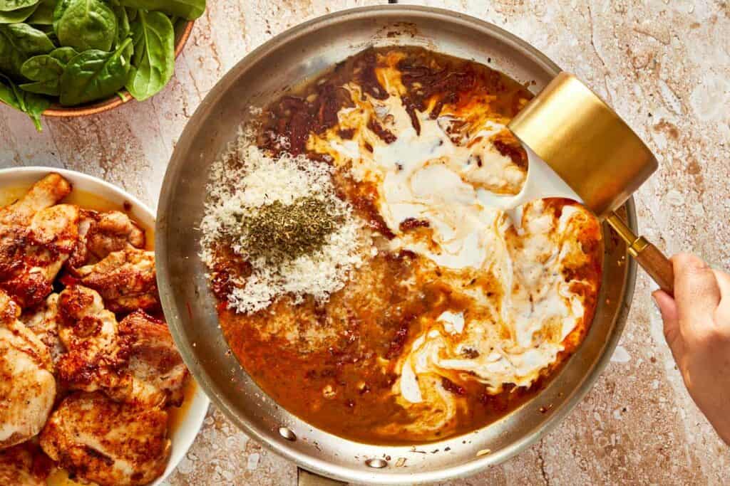 a top down shot of a metal bowl filled with red sauce and spices and also a cup of white cream being poured into it