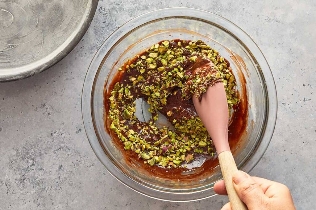 placing crushed pistachios in a glass bowl with chocolate using a wooden spoon in a large glass bowl.