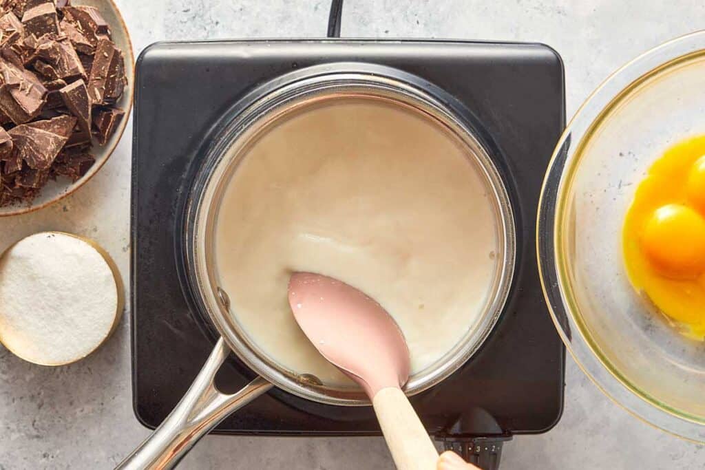 cream in a saucepan being mixed with a wooden spoon on a stove top, top down view