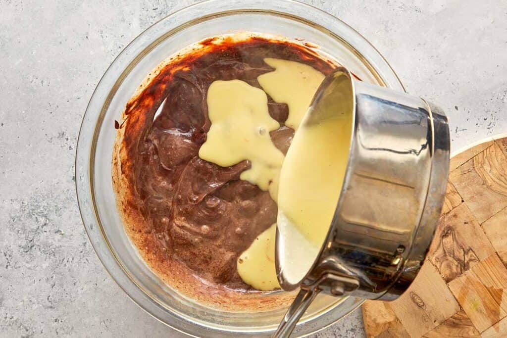 the remainder of the custard from a pot being poured into a glass bowl with chocolate inside