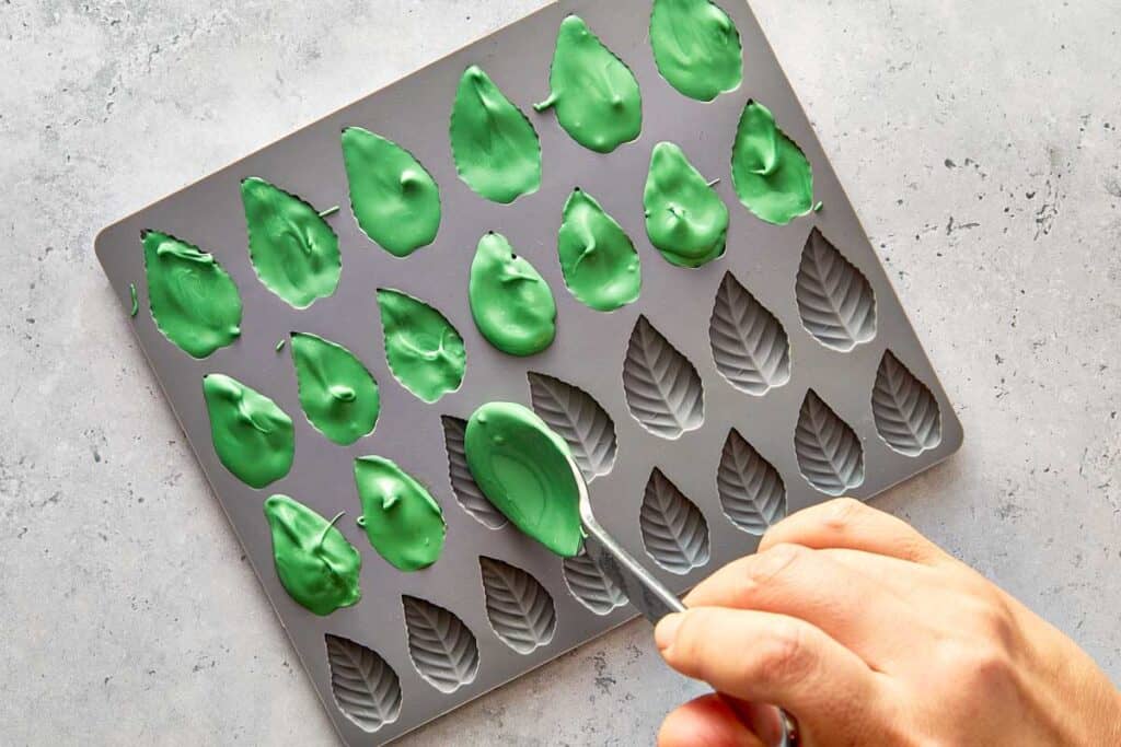 creating green chocolate leaves by placing green chocolate in a leaf shaped silicone mould tray there are rows of leaves 6 x 4 using a teaspoon to place the chocolate