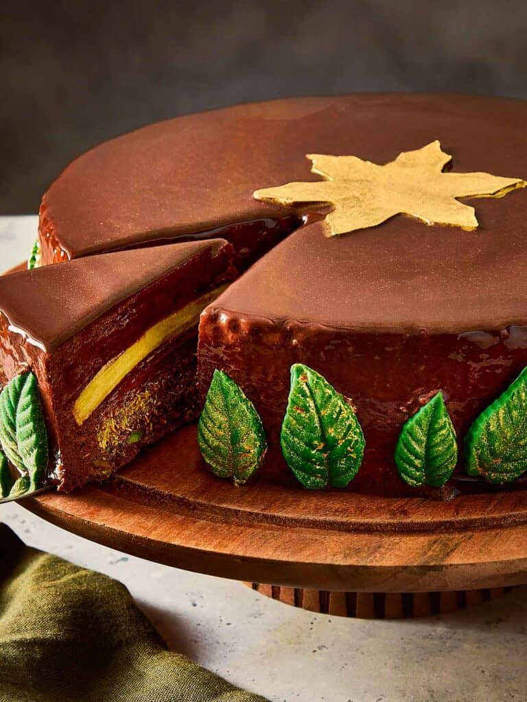 a round glazed chocolate cake side view sitting on a cake tray decorated with green leaves and a golden star on top with a slice cut out showing the layers