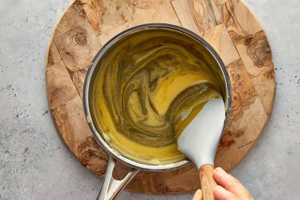 mixing pistachio paste in a bowl of custard off heat resting on a wooden board.