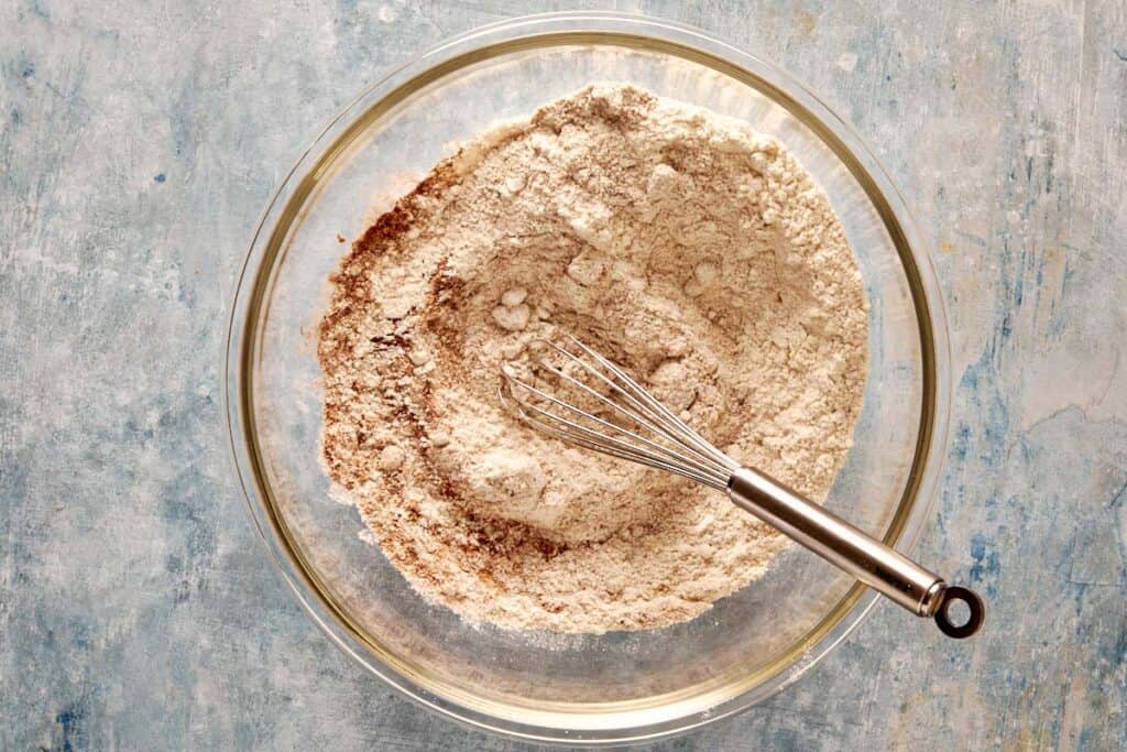 a top down view of a glass bowl with all the dry ingredients mixed with a metal whisk which is currently resting on the side of the bowl. - 4
