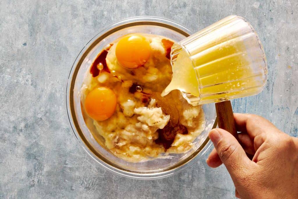 a top down view of a glass bowl with wet ingredients being mixed into the dry ingredients and a glass jug of liquid being poured into the bowl. - 5