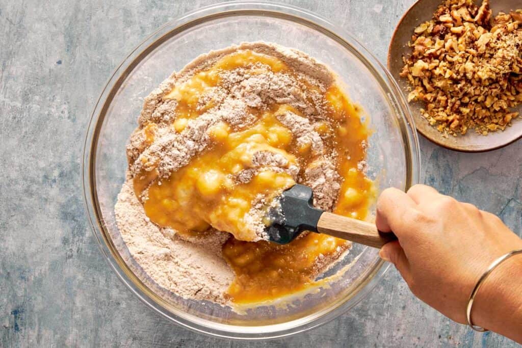 a top down view of a green spatula mixing both the wet ingredients and dry ingredients in a glass bowl sitting on top of a bench - 7