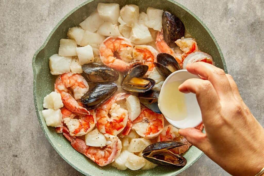 a top down view of seafood in a pan being cooked with what looks like white wine being poured into the pan. - 8