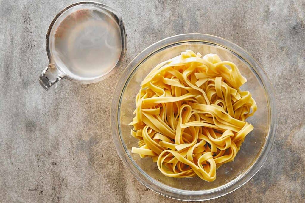 a top down view of a glass bowl full of cooked pasta and a jug of water next to it. - 11