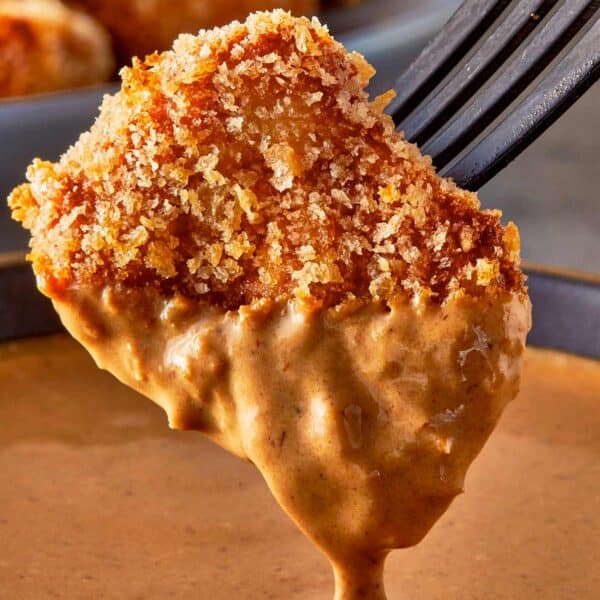 a side view of a single chicken nugget half dipped in sauce dripping hovering over a sauce bowl, held up by a fork