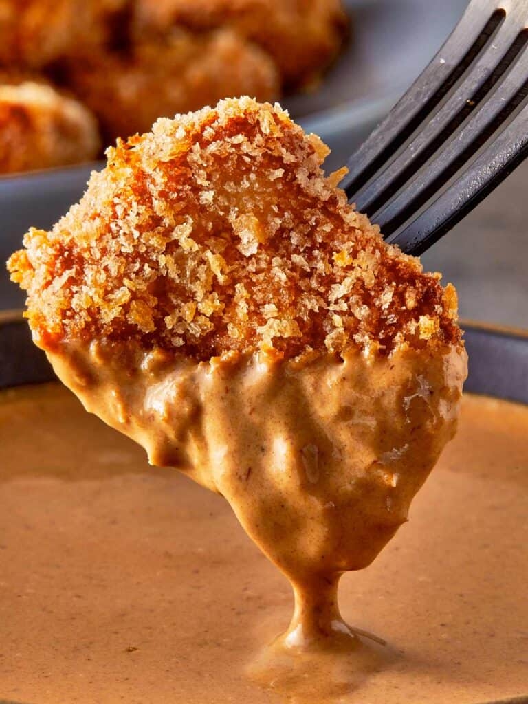 a side view of a single chicken nugget half dipped in sauce dripping hovering over a sauce bowl, held up by a fork