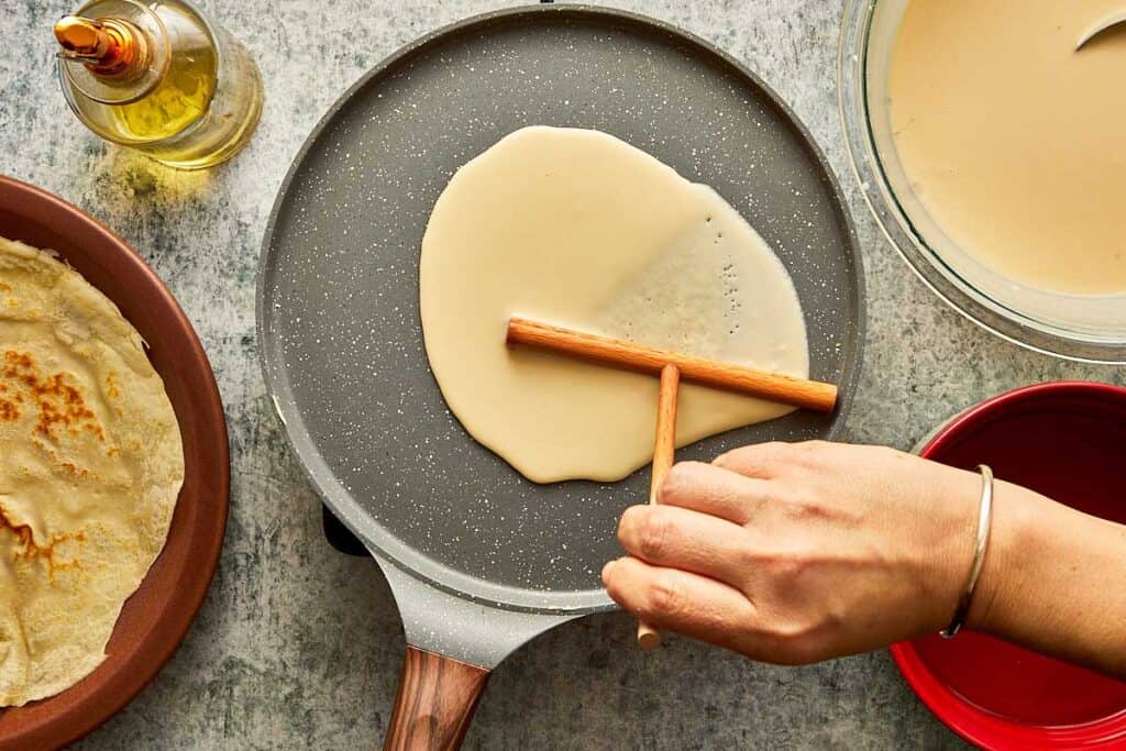 a top down view of a crepe being made using a crepe tool to flatten the batter on top of a crepe pan - 16
