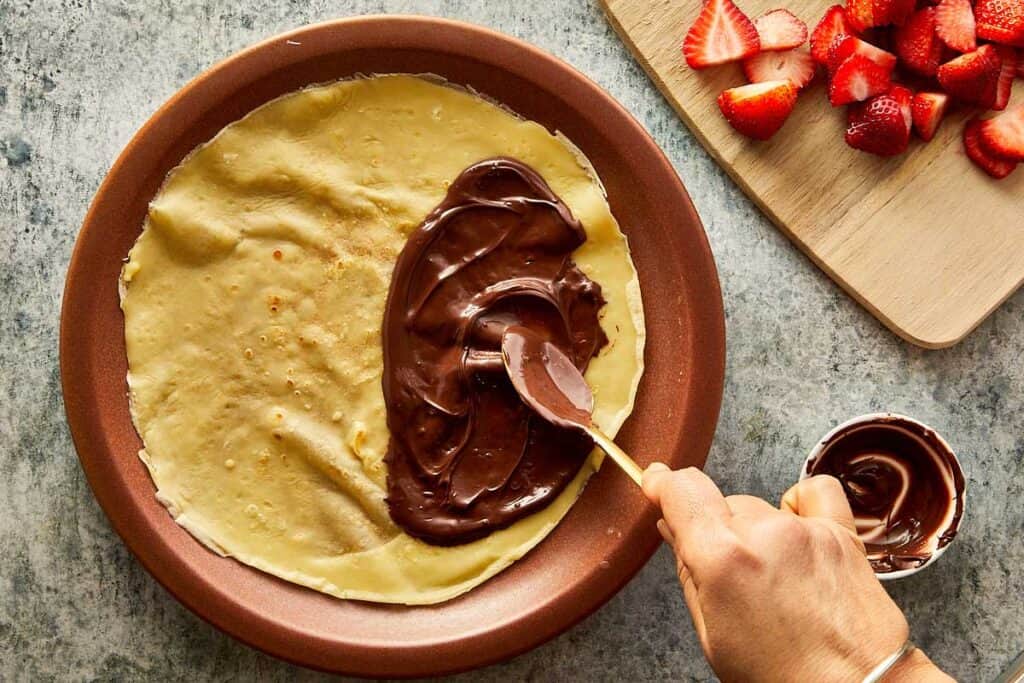 a cooked crepe sitting on top of a ceramic brown plate, the crepe is creased as it sits on the plate and a golden spoon is covering one side of the crepe with Nutella paste - 21