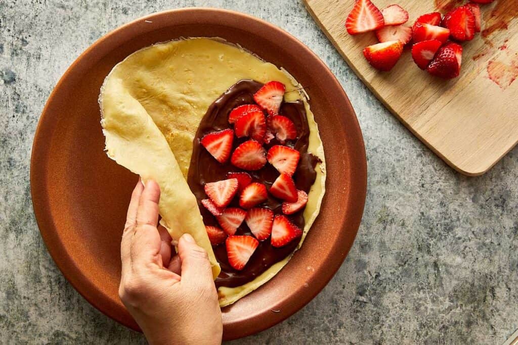 a cooked crepe sitting on top of a ceramic brown plate, the crepe is creased as it sits on the plate with nutella paste on one side covered with strawberries on top which have been quartered - 22