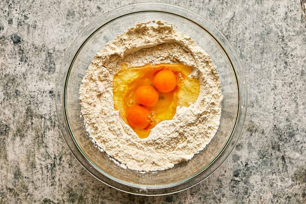 a top down view of a glass mixing bowl on a kitchen bench with 3 eggs in the middle of a flour mix - 7