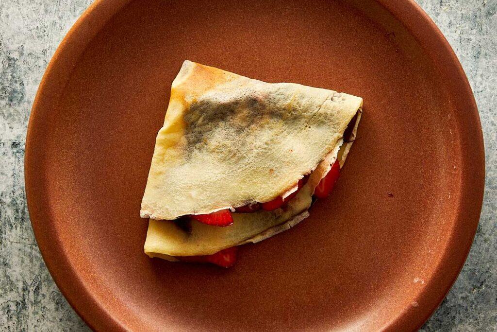 a top down view of a brown ceramic plate with a folded crepe filled with nutella and strawberries. - 23