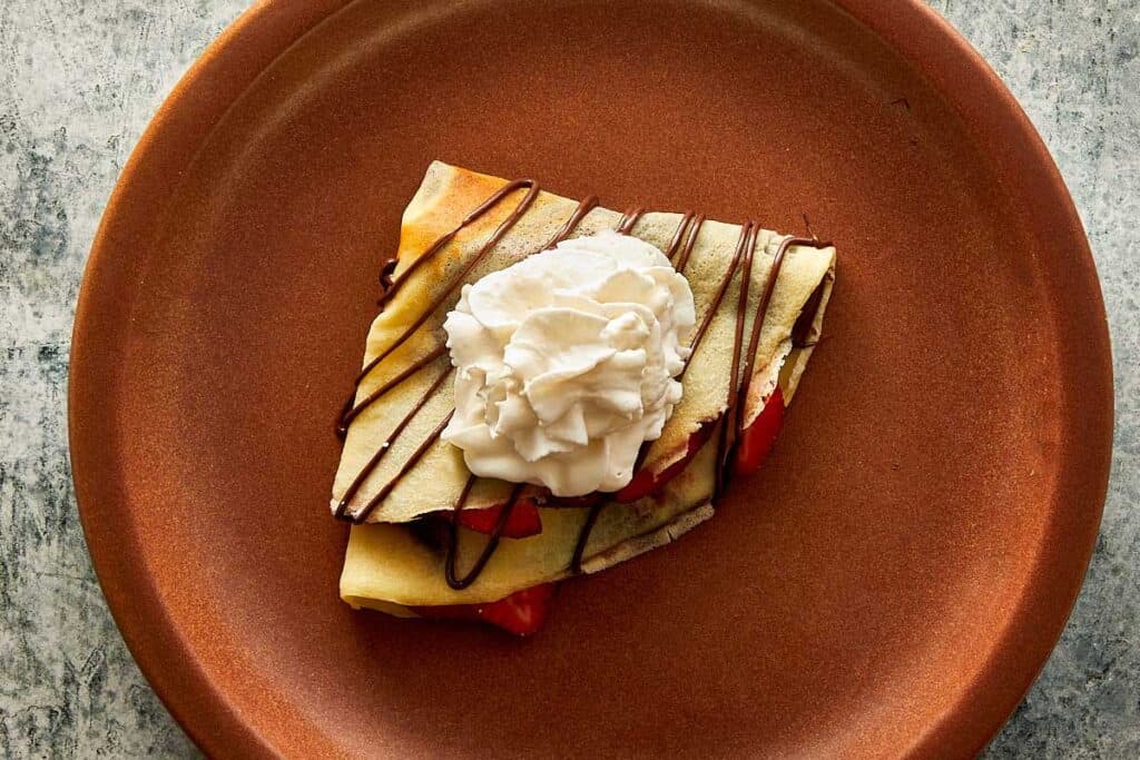 a top down view of a ceramic brown plate with a quartered crepe sitting on top with a blob of cream and some drizzle zig zagged topping of Nutella - 24