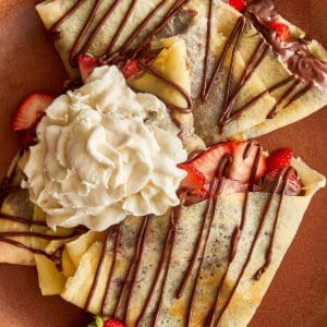 a top down view of a ceramic brown plate with a quartered crepe sitting on top with a blob of cream and some drizzle zig zagged topping of Nutella 3 of them on a plate with strawberry quarters on top