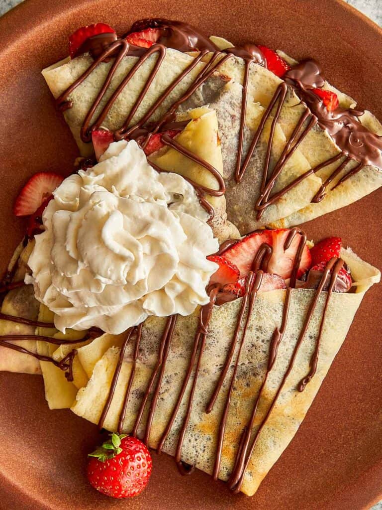 a top down view of a ceramic brown plate with a quartered crepe sitting on top with a blob of cream and some drizzle zig zagged topping of Nutella 3 of them on a plate with strawberry quarters on top