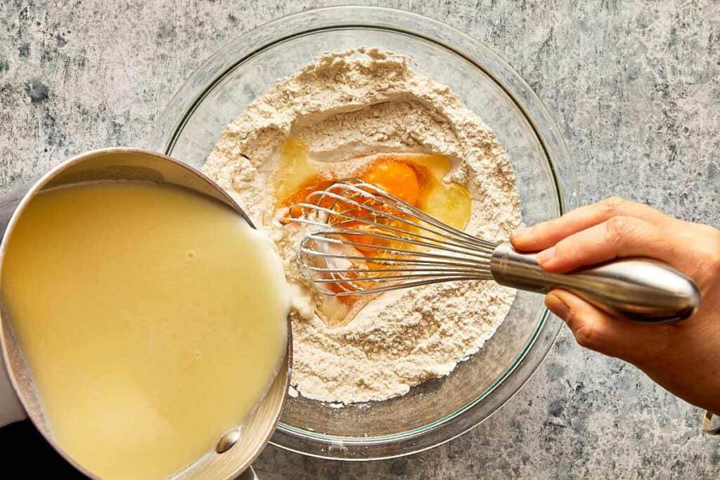 a top down view of a glass mixing bowl on a bench filled with flour and egg mixture and a bowl of milk being poured into it slowly while a hand is whisking everything together. - 8