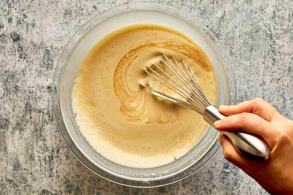 a top down view of a glass bowl and a whisk being held by a female hand mixing a thick white mixture which is getting smooth - 10