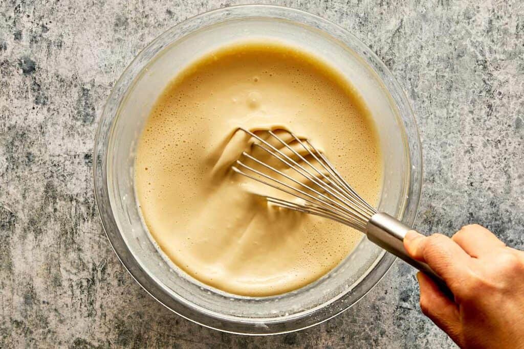 a top down view of a glass bowl and a whisk being held by a female hand mixing a thick white mixture which is smooth - 11
