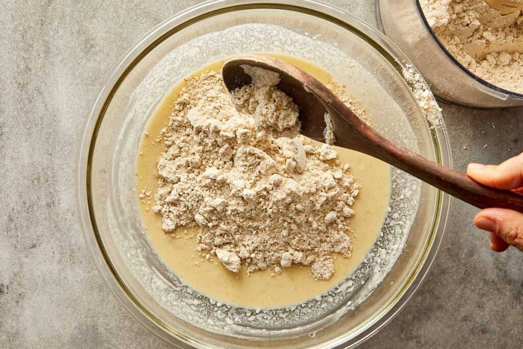 a top down image of a glass mixing bowl with white batter in the bottom of the bowl and white powder on top getting ready to be mixed with a wooden mixing spoon - 8