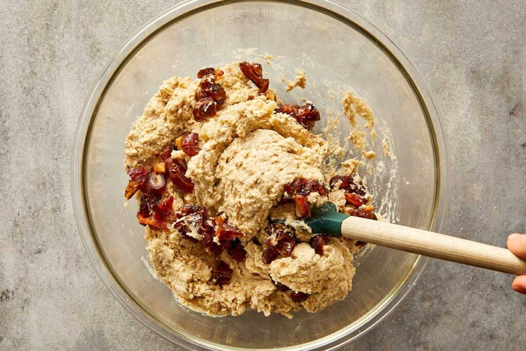 a top down image of a glass mixing bowl with white batter in the bottom of the bowl and white powder on top getting ready to be mixed with a wooden mixing spoon with fruit inside - 9