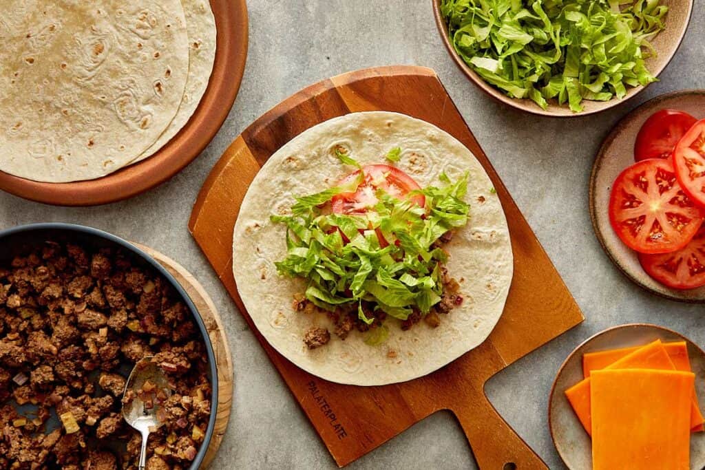 a top down view of a wrap on top of a wooden board sitting on a kitchen bench, the wrap has cooked mince, slice of cheese, lettuce and tomatoes - 12