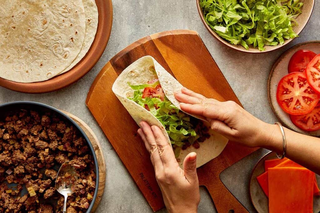 a top down view of a wrap being made by two female hands, the wrap is filled with mince meat and lettuce and its on top of a wooden board. - 13
