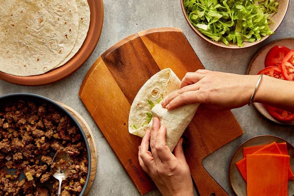 a top down view of a wrap being made by two female hands, the wrap is filled with mince meat and lettuce and its on top of a wooden board. - 15