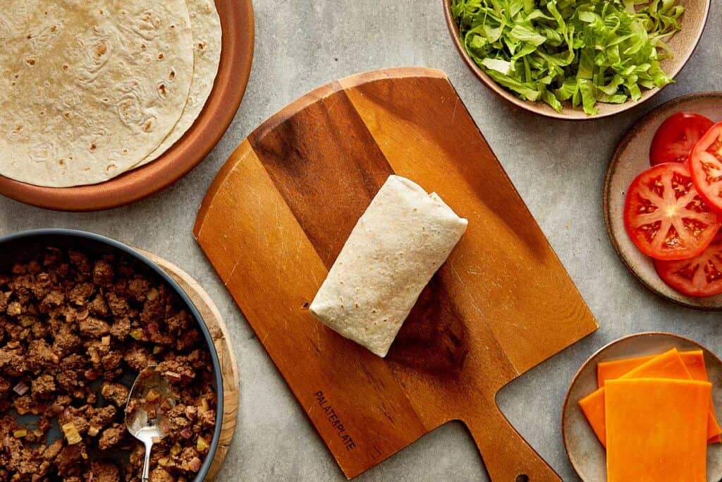 a top down view of a wrap being made by two female hands, the wrap is filled with mince meat and lettuce and its on top of a wooden board. - 16