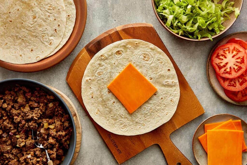 a top down view of a wrap on top of a wooden board sitting on a kitchen bench, the wrap has a slice of cheese on it. - 10